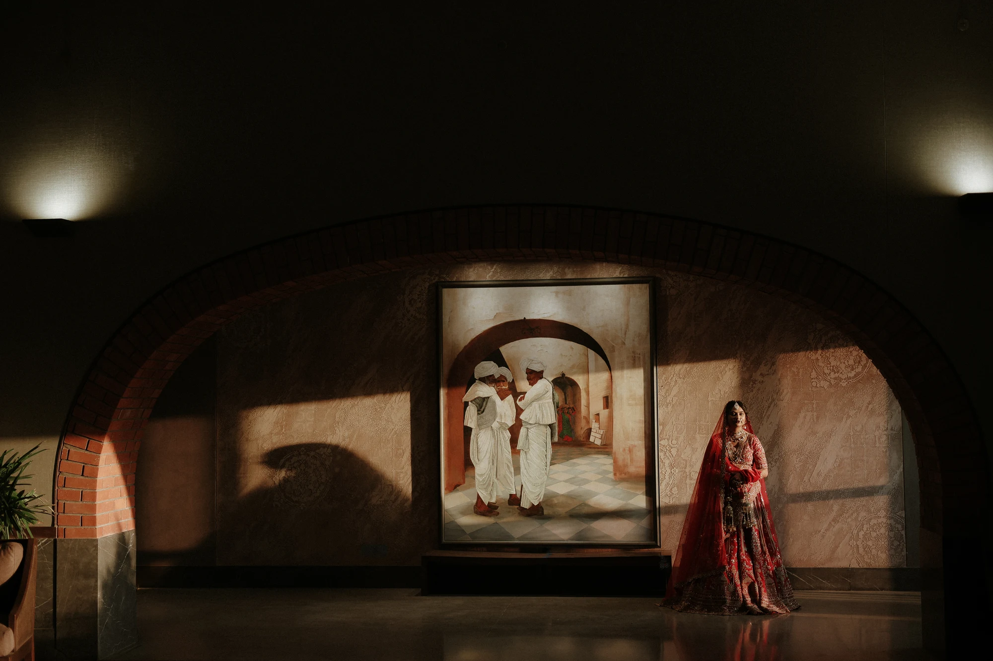 Regal bride in crimson lehenga standing under sunlit architectural archway